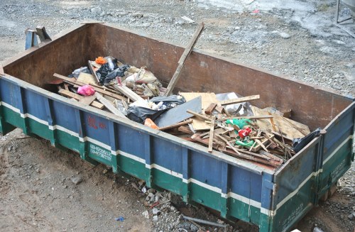 A large, rectangular metal skip situated outdoors on a patch of gravelly ground, filled with various scattered debris including wooden planks, plastic bags, cardboard, and loose rubbish. The skip’s side panels are painted blue with green and white horizontal stripes, and the interior shows rust and wear indicating frequent use. The debris appears partially collapsed and disorganized, with some items leaning against the sides of the skip and others piled loosely within. Surrounding the skip is uneven ground with small stones and gravel, suggesting a construction or commercial waste collection site. There are no other objects or structures visible in the immediate background. This image relates to rubbish removal services provided by Commercial Waste Brent Cross, serving areas including Brent Cross and nearby parts of northwest London, emphasizing waste collection for commercial or construction purposes.