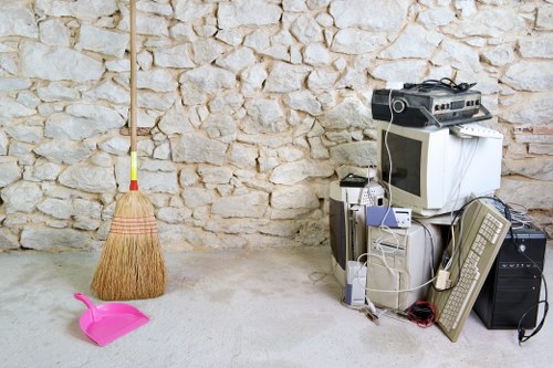 The image shows a collection of old electronic equipment and miscellaneous items placed on the floor next to a stone wall in an indoor or outdoor setting, possibly a storage or utility area. The items include a CRT computer monitor, an old desktop computer tower, a beige keyboard, and a variety of tangled cables and smaller electronic devices. To the left, leaning against the wall, is a wooden broom with a natural bristle head and a pastel pink dustpan. The floor appears to be concrete or similar material, and the scene is lit evenly, indicating ambient lighting. This arrangement of discarded electronics and cleaning tools is typical of rubbish or waste awaiting removal, aligning with services offered by Commercial Waste Brent Cross in the London borough. The setting suggests a cluttered space where unwanted electronic waste and cleaning equipment are stored temporarily before disposal or recycling, contextualising the rubbish removal process in the Brent Cross area, close to NW4 postcode.