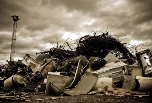 A large pile of discarded household appliances and metal debris, including washing machines, metal cabinets, and various scrap metal parts, are stacked outdoors on a barren, earth surface. The objects appear weathered and rusted, with some items leaning against each other or lying partially on their sides. The background features a cloudy sky with dark, brooding clouds, and a tall metal tower is visible on the left side of the image, suggesting an industrial or outdoor storage area near Brent Cross. This scene illustrates the type of bulky waste that Commercial Waste Brent Cross may collect and remove as part of their rubbish disposal services, emphasizing the need for proper waste management for large, metal household items before disposal or recycling.