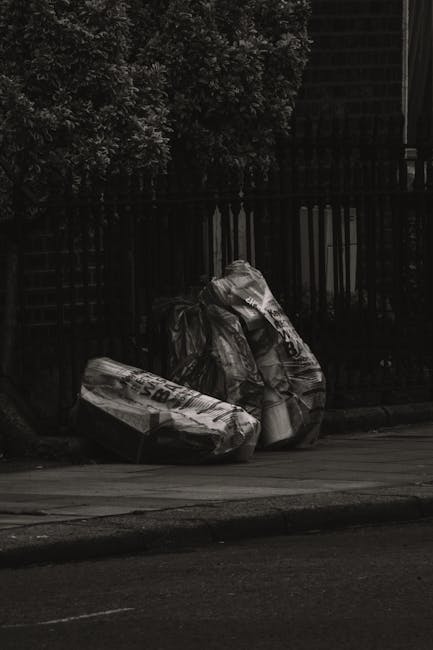 A narrow urban alleyway cluttered with a large, black, weathered recycling or waste bag in the foreground, positioned on a concrete surface that shows signs of dirt and wear. Behind the bag, there is 